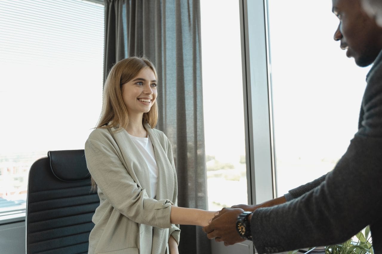 who-we-are Woman smiling and shaking hands at a business office, signaling a successful job interview.
