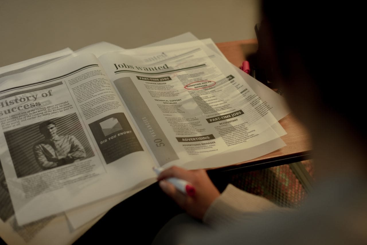 team-02 Person reviewing job listings in a newspaper, holding a pink marker for highlighting job ads.